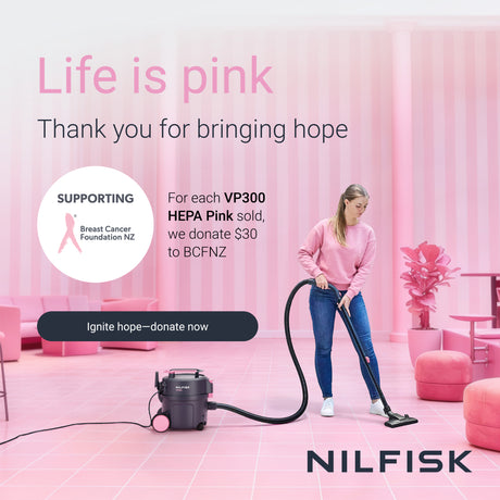 A woman in a pink sweatshirt and blue jeans uses a Nilfisk HEPA Pink vacuum cleaner in a pink room. The image includes promotional text for Breast Cancer Foundation NZ and the Nilfisk brand.