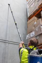 Person in a warehouse using a telescopic tool to reach high shelves with stacked boxes.