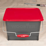 A gray Wooster paint bucket with a red lid and red handle grip, shown on a beige fabric background.
