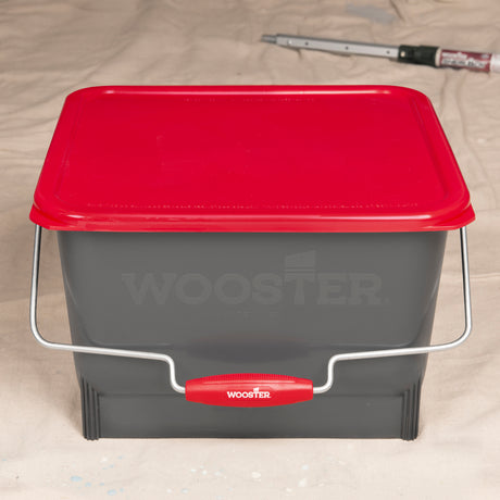 A gray Wooster paint bucket with a red lid and red handle grip, shown on a beige fabric background.