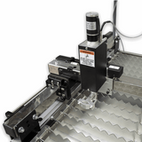 Industrial laser marking machine with a black and silver Z-axis gantry system, featuring a warning label and a metallic zig-zag bed, shown against a white background.