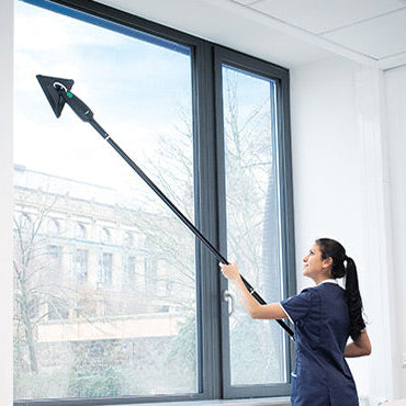 A woman in a navy scrub top uses a stingray extension pole  window cleaning tool with a triangular clean a large office window.
